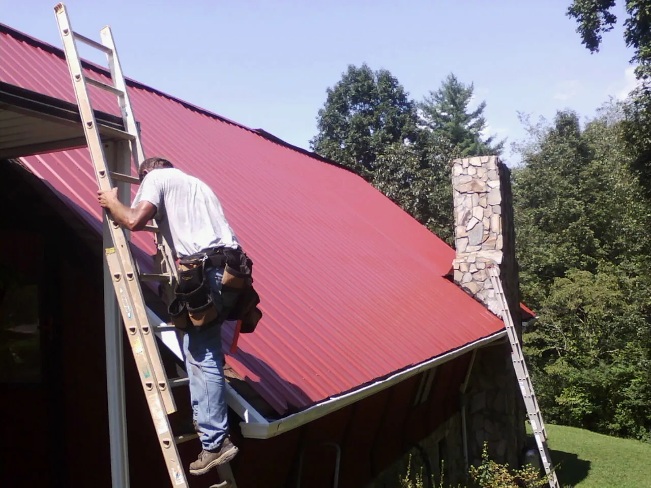Roofer on a ladder beside a red metal roof and stone chimney — Soffit & Fascia Repair by Roofing Brothers in Castalia